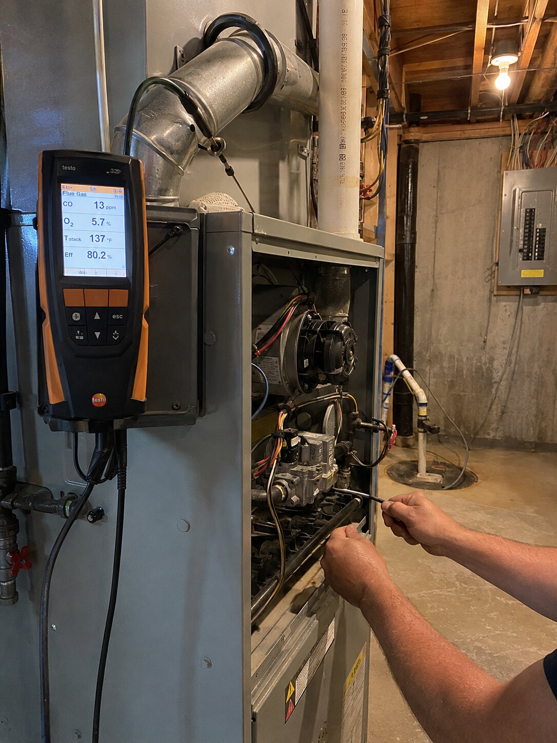 Combustion analyzer measuring CO and O2 levels in residential gas furnace flue with technician adjusting manifold pressure